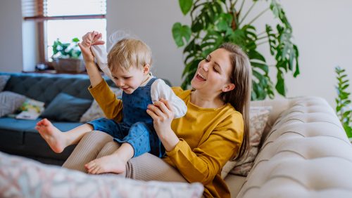 ouders-kind-opvoeden A young cheerful mother playing with her little child and having fun when lifting her up on sofa.
